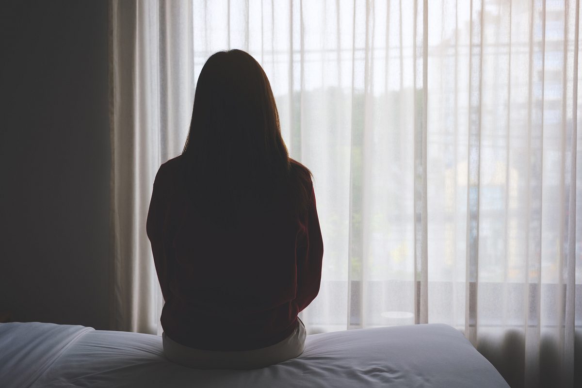 Frau sitzt mit dem Rücken zum Betrachter auf dem Bett und schaut aus dem Fenster (Symbolbild).