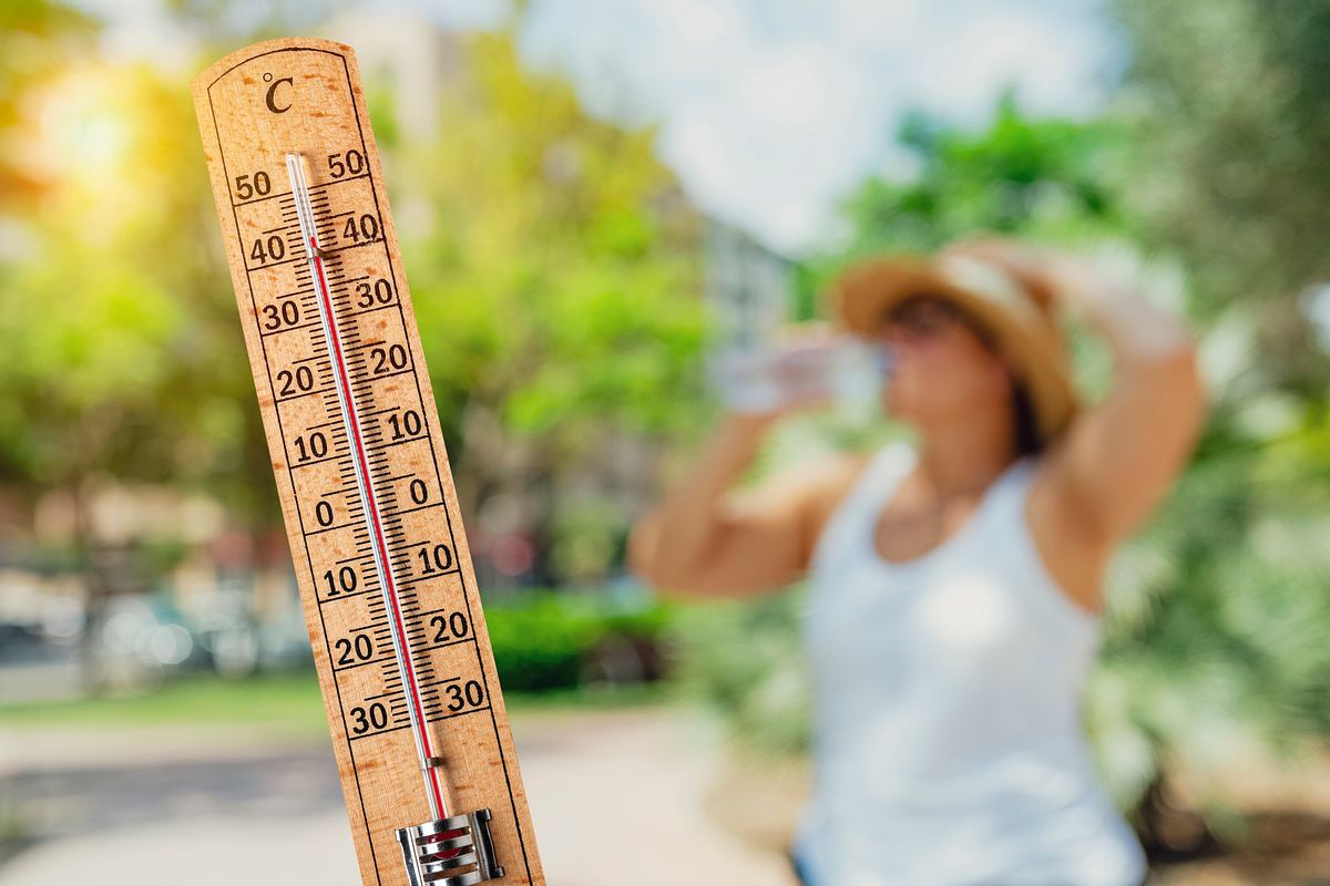 Thermometer, das über 40 Grad anzeigt - im Hintergrund trinkt eine Frau mit Sonnenhut Wasser.