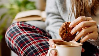 Nahaufnahme einer unerkennbaren Frau, die zu Hause auf dem Sofa Kaffee trinkt und Kekse isst. - Foto: Anna Ostanina/iStock