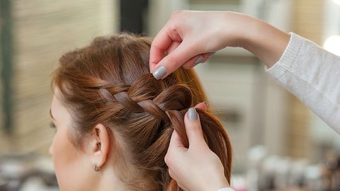 Frau mit rötlichem Haar bekommt seitlichen Zopf gelfochten. - Foto: dimid_86/iStock (iStock)