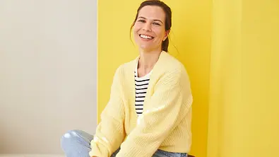 Ein Frau in Jeans, Streifenshirt und gelbem Cardigan sitzend, ein Bein übergeschlagen - Foto: Almased