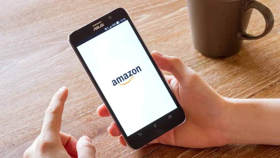 Amazon ändert Retouren-Regeln Ein Mann sitzt an einem Holztisch und hält ein Smartphone in der Hand. Auf dem Display ist die geöffnete Amazon App zu sehen.
