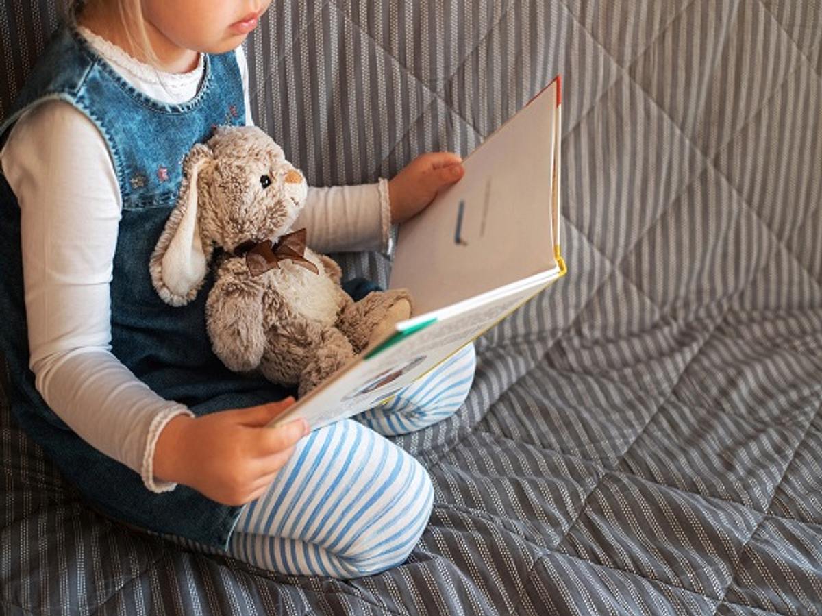 Kleines Mädchen mit Kuscheltier liest Buch auf dem Sofa