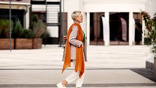 Eine blonde, weiblich gelesene Person flaniert durch die Stadt und trägt Sonnenbrille, Karo-Blazer, Loafers und einen orangenen Schal - Foto: iStock/ AnnaZhuk