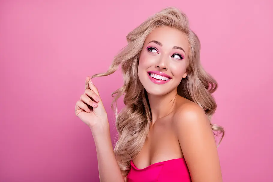 Barbie-Droge Melanotan Blonde Frau im Barbie-Look spielt vor rosa Hintergrund mit ihren Haaren und blickt verträumt zur Seite.