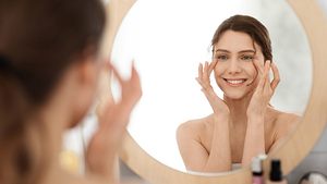Frau lächelt in den Spiegel und trägt sich Gesichtspflege auf - Foto: iStock / Prostock-Studio