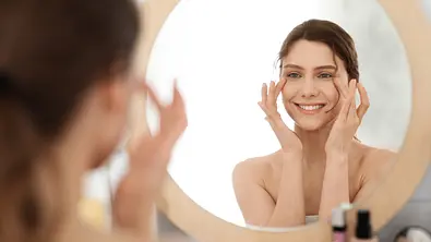 Frau lächelt in den Spiegel und trägt sich Gesichtspflege auf - Foto: iStock / Prostock-Studio