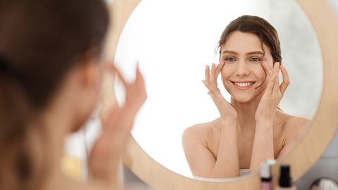 Frau lächelt in den Spiegel und trägt sich Gesichtspflege auf - Foto: iStock / Prostock-Studio