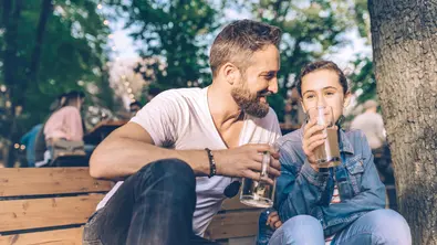 Vater sitzt mit Teenager-Tochter auf einer Bank im Biergarten und trinkt Bier. - Foto: iStock/golero