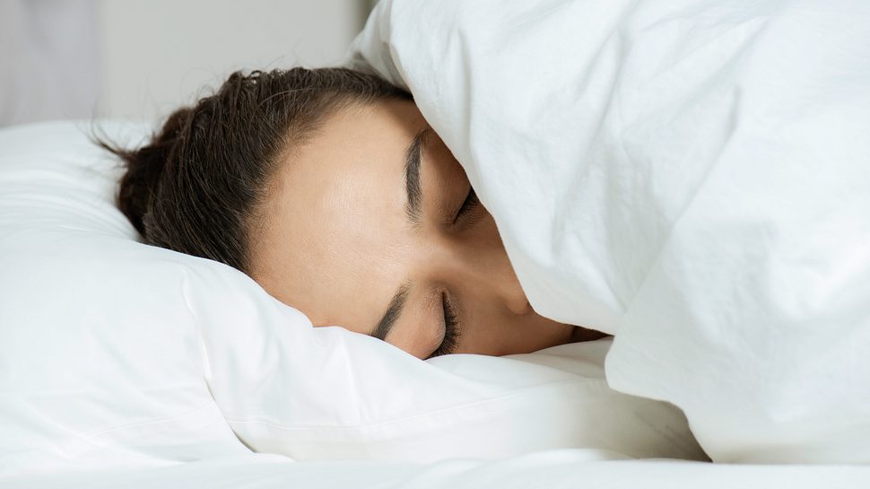Eine schlafende kaukasische Frau im Bett versteckt ihr Gesicht unter der Decke. - Foto: simarik/iStock