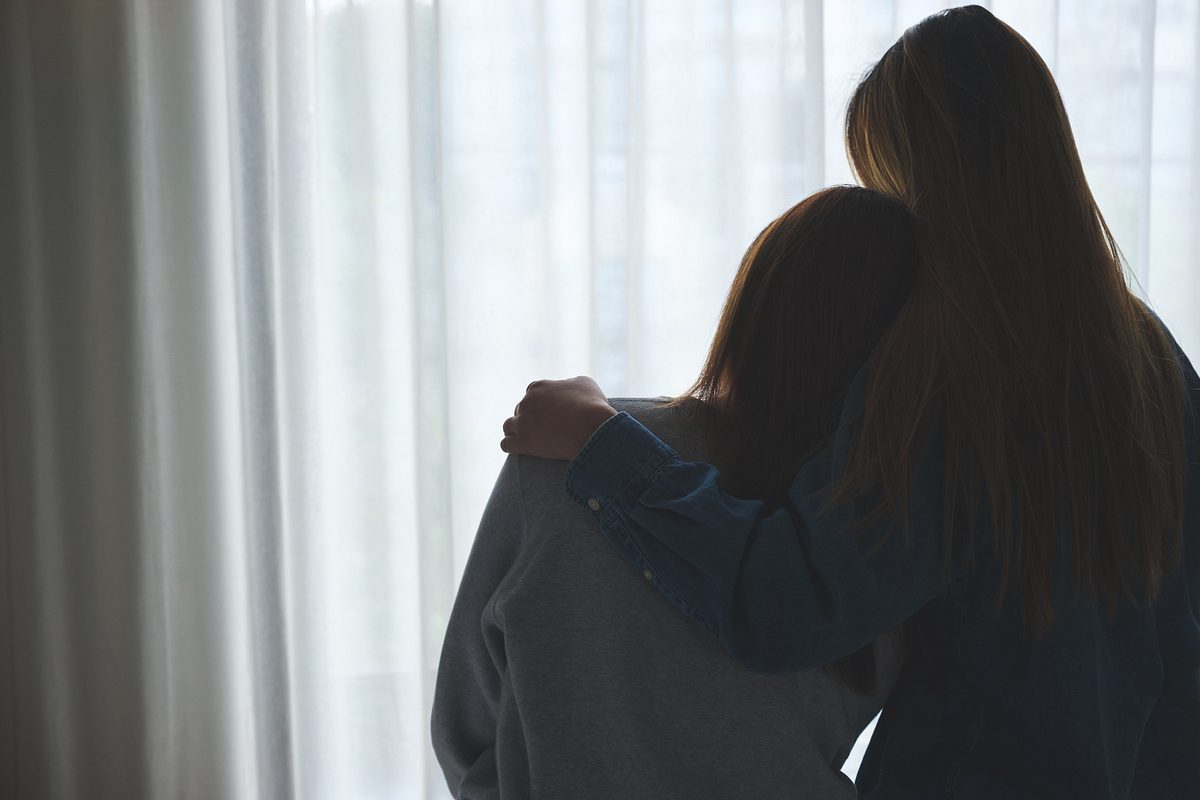 Zwei Frauen stehen Arm in Arm am Fenster und blicken nach draußen.