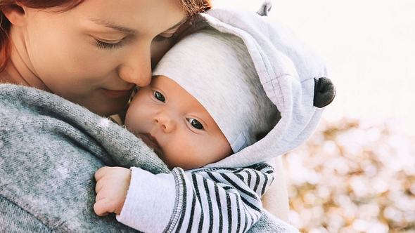 Mama mit Baby auf dem Arm, das mit Mütze und warmen Jäckchen abgebildet ist - Foto: iStock/NataliaDeriabina