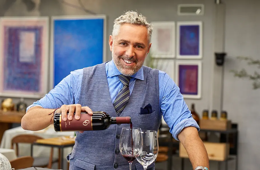 First Dates Hotel Roland Trettl steht in einem Restaurant und schenkt Rotwein in zwei Weingläser ein; er trägt Hemd, Krawatte und Weste, im Hintergrund sind Tische und gerahmte Bilder an der Wand zu sehen.