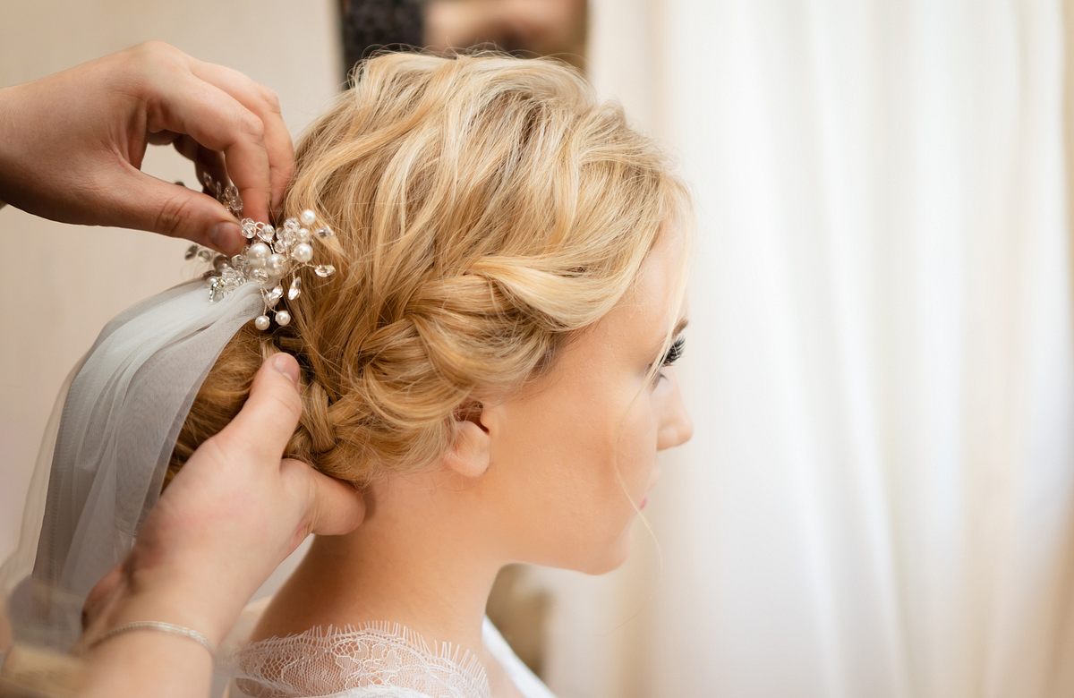 Blonde Braut trägt geflochtene Hochzeitsfrisur mit Schleier und Haarnadel.