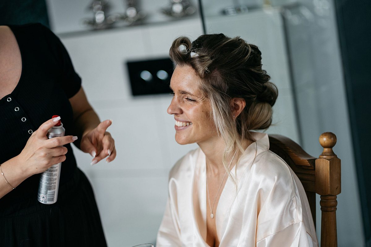 Eine Braut im Bademantel hat Lockenwickler im Haar und wird von einer zweiten Frau mit Haarspray in der Hand für ihre Hochzeit gestylt.