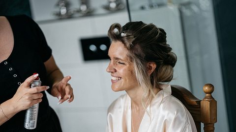 Eine Braut im Bademantel hat Lockenwickler im Haar und wird von einer zweiten Frau mit Haarspray in der Hand für ihre Hochzeit gestylt. - Foto: fotojuwelier/iStock (Themenbild)