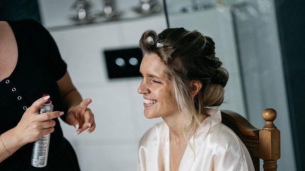 Eine Braut im Bademantel hat Lockenwickler im Haar und wird von einer zweiten Frau mit Haarspray in der Hand für ihre Hochzeit gestylt. - Foto: fotojuwelier/iStock (Themenbild)