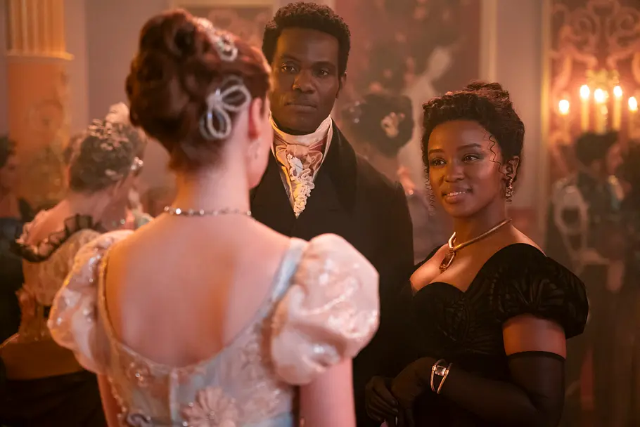 Francesca (Hannah Dodd), John (Victor Alli) und Michaela (Masali Baduza). Ein Mann und eine Frau im eleganten Regency-Stil schauen auf eine Frau in einem cremefarbenen Kleid, die ihnen den Rücken zukehrt. Die Szene spielt auf einem rauschenden Ball mit festlich gekleideten Gästen und Kerzenlicht im Hintergrund. 