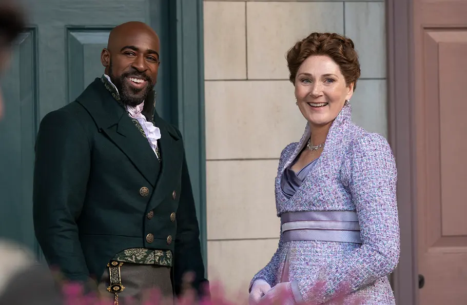 Lady Violet Bridgerton in einem hellvioletten Kleid und Lord Anderson im dunklen Gehrock stehen nebeneinander vor einer Haustür und lächeln freundlich in die Kamera; im Vordergrund sind verschwommen Blumen zu sehen.