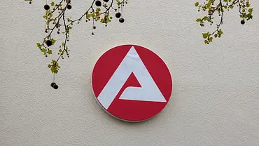 Logo der Bundesagentur für Arbeit an einer Außenwand, darüber Äste mit Blättern. - Foto: IMAGO / NurPhoto Logo der Bundesagentur für Arbeit an einer Außenwand, darüber Äste mit Blättern. - Foto: IMAGO / NurPhoto