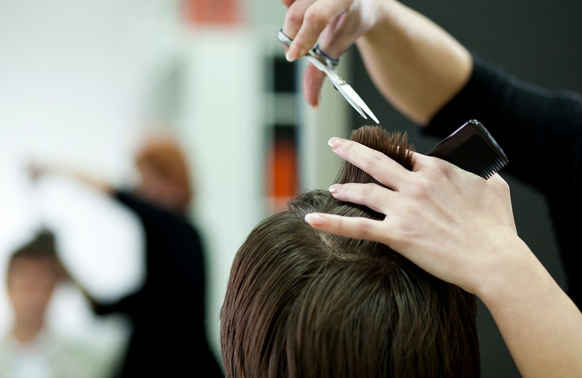 Linke Hand mit Kamm hält kurze Haare hoch, rechte Hand mit Schere schneidet ins Haar.