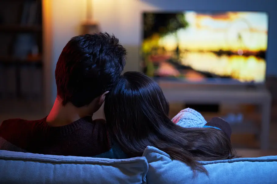 Was wir aus Liebesfilmen lernen können Rückenansicht eines heterosexuellen Paares, das zuhause auf dem Sofa einen Film schaut. Er legt einen Arm um ihre Schulter.