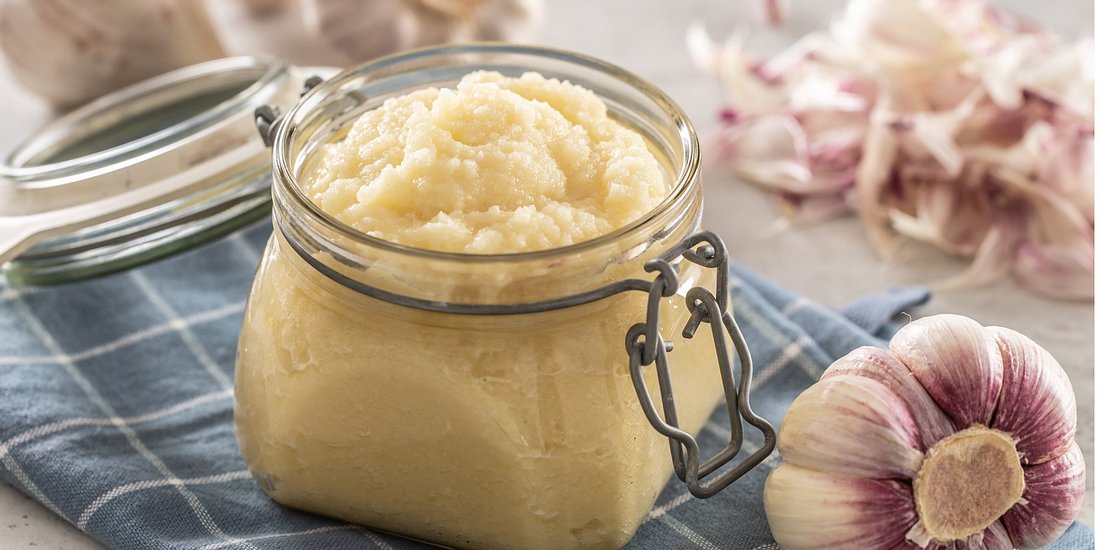 Aromatic garlic paste in a glass jar laid on rustic kitchen cloth with bulbs and peeled cloves.