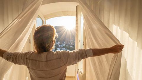 Frau öffnet Vorhänge am Fenster (Themenbild) - Foto: Mystockimages/iStock