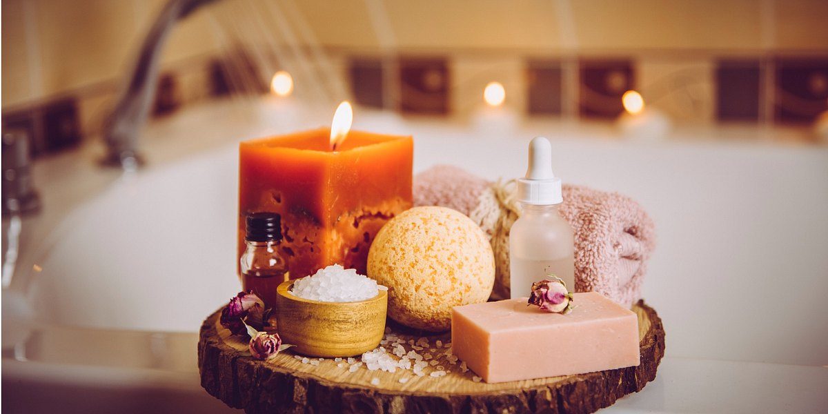 Home spa products on wooden tray: bar of soap, bath bomb, aroma bath salt, essential and massage oils, candle burning, rolled towel inside bathroom by tub running water.