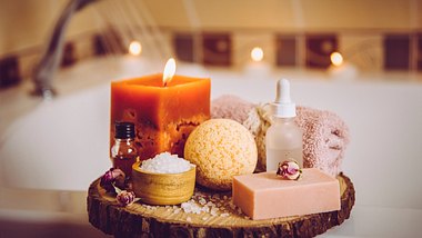 Home spa products on wooden tray: bar of soap, bath bomb, aroma bath salt, essential and massage oils, candle burning, rolled towel inside bathroom by tub running water. - Foto: Helin Loik-Tomson/iStock