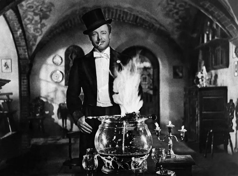 „Die Feuerzangenbowle“ Ein Mann im Frack und Zylinder steht lächelnd in einem altmodisch eingerichteten Raum hinter einer großen Glasschale, aus der eine Flamme emporsteigt.