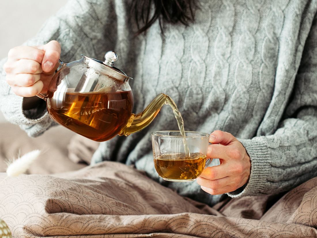 Frau in Strickpullover gießt heißen Tee aus der Glasteekanne in die Tasse