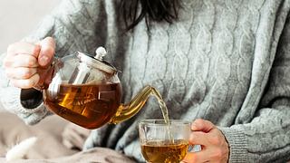 Frau in Strickpullover gießt heißen Tee aus der Glasteekanne in die Tasse - Foto: photoguns/iStock