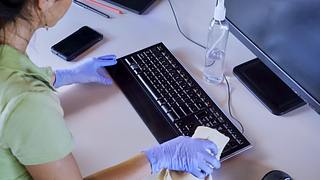 Eine Frau mit Latex-Handschuhen reinigt die Tastatur ihres Computers - Foto: iStock/apomares