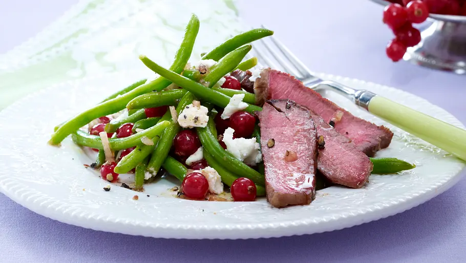 Eiweiß-Gericht: Bohnen mit Ziegenkäse Grüne Bohnen, Johannisbeeren, zerbröselter Ziegenkäse und drei Streifen Rinderhüftsteak auf einem weißen Teller serviert.