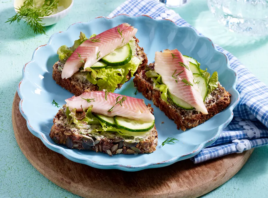 Eiweißreiches Rezept: Forellen-Eiweißbrot 3 Scheiben Körnerbrot mit Gurke, Salat und Forelle auf einem blauen Teller.