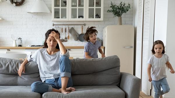 Kinder rennen um Sofa, Mutter sitzt genervt darauf (Themenbild) - Foto: fizkes/iStock