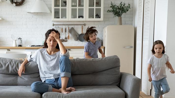 Kinder rennen um Sofa, Mutter sitzt genervt darauf (Themenbild) - Foto: fizkes/iStock