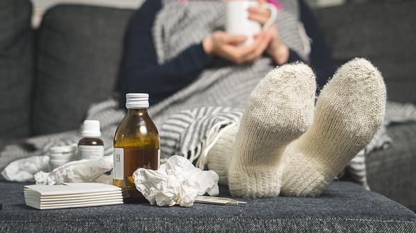 Frau liegt neben benutzten Taschentüchern und Medikamentenfläschchen in Wollsocken auf dem Sofa und hat eine Tasse Tee in der Hand (Themenbild) - Foto: Tero Vesalainen/iStock