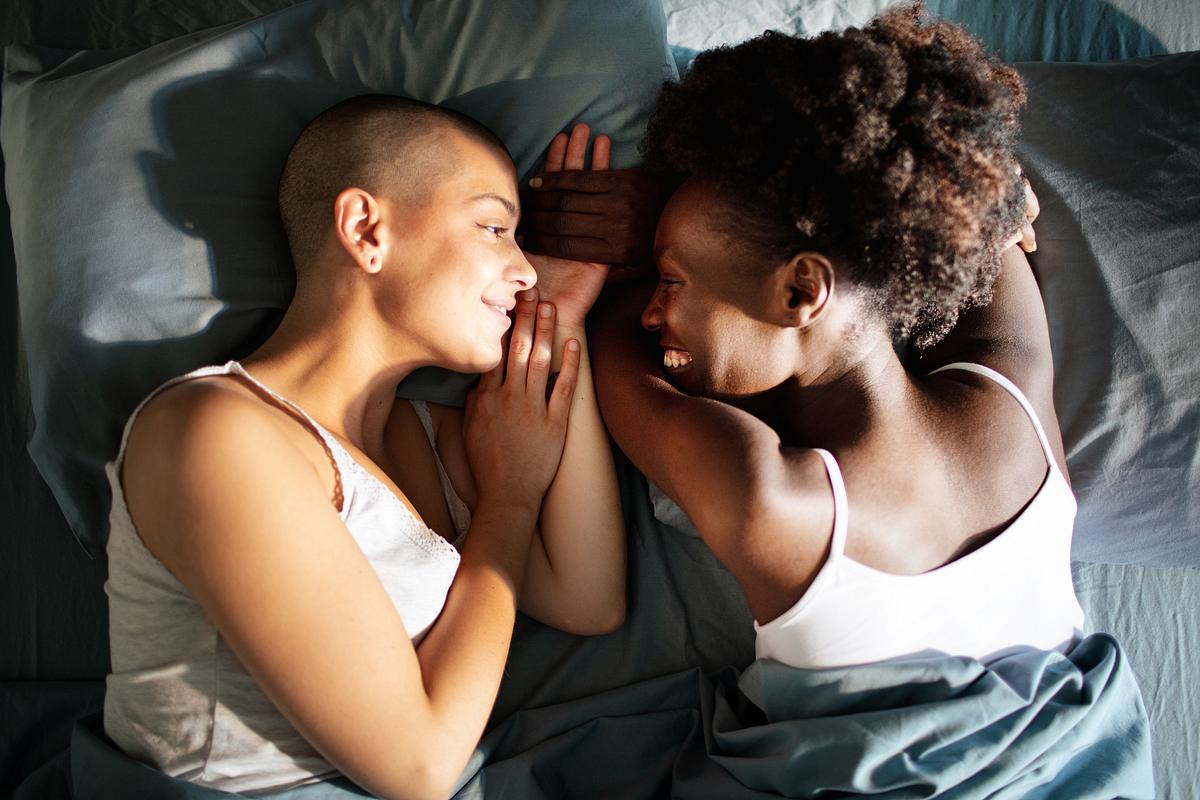 Zwei Frauen liegen im Bett und teilen einen zärtlichen Moment. Sie halten sich an der Hand, sehen sich in die Augen und lachen.