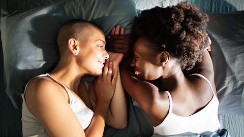 Zwei Frauen liegen im Bett und teilen einen zärtlichen Moment. Sie halten sich an der Hand, sehen sich in die Augen und lachen. - Foto: iStock/ vorDa 