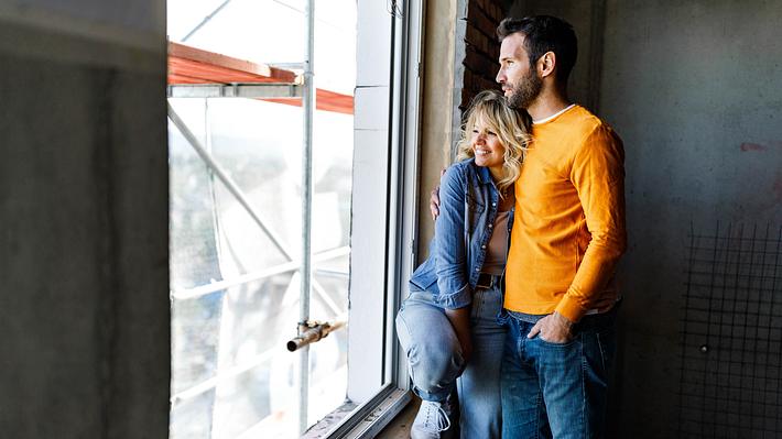 Ein glückliches Paar steht am Fenster einer Baustelle und beobachtet den Sonnenaufgang. - Foto: skynesher / iStock