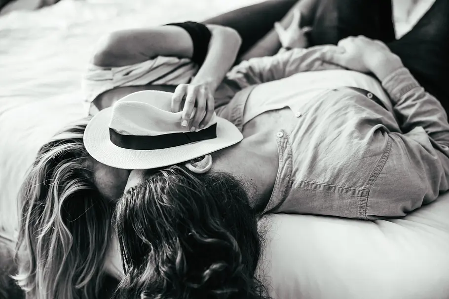 Erotische Liebesgeschichte Schwarz weiß Foto eines verliebten Paares, das auf einem Bett liegt und sich küsst. Sie hält einen Hut vor ihre Gesichter.