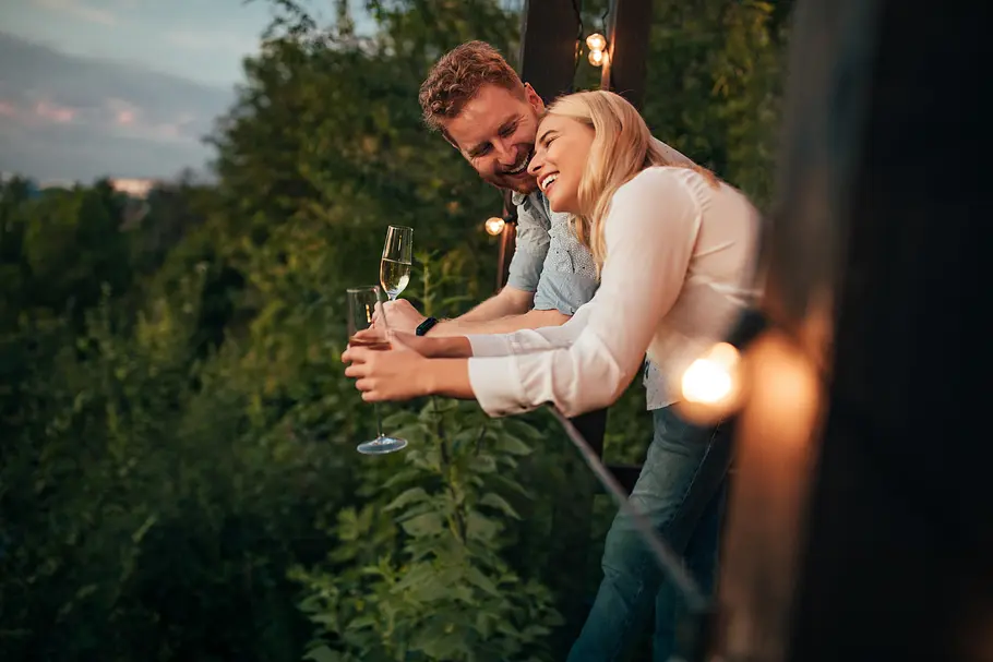 Erotische Liebesgeschichte Ein verliebtes heterosexuelles Paar steht auf einem Balkon mit einem Ausblick auf den Wald, trinkt ein Glas Champagner, redet und lacht.