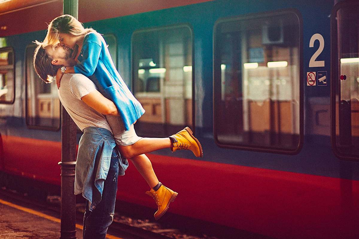 Ein verliebtes Paar umarmt sich küssend am Bahngleis, erhebt sie hoch und hinter ihnen steht ein Zug. 