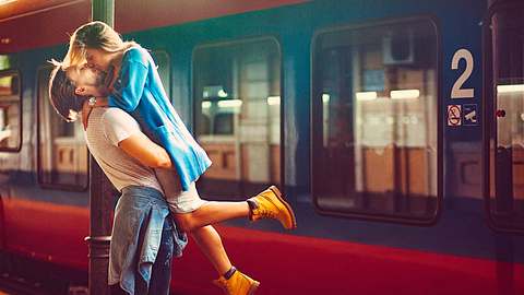 Ein verliebtes Paar umarmt sich küssend am Bahngleis, erhebt sie hoch und hinter ihnen steht ein Zug.  - Foto: gruizza/iStock