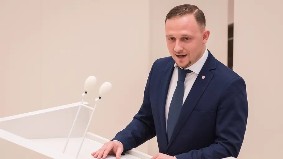 Felix Teichner Felix Teichner steht in einem blauen Anzug vor einem weißen Pult und spricht.