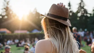 Frau mit blondem Haar und Hut steht vor einer Wiese mit Menschen. - Foto: Halfpoint/iStock (Themenbild)