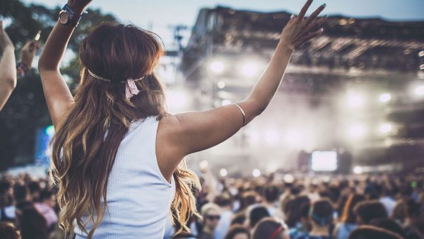 Eine Frau mit gelocktem Haar steht vor einer Konzertbühne und reckt die Arme in die Luft.  - Foto: skynesher/iStock (Themenbild)