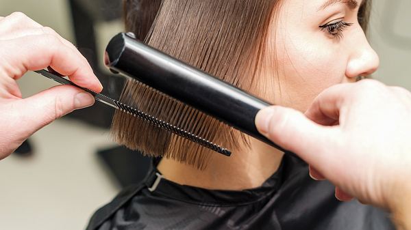 Frau mit braunen Haaren bekommt die Haare mit einem Glätteisen frisiert. - Foto: okskukuruza/iStock (Themenbild)